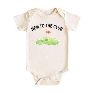 May include: Cream-colored baby onesie with the text "NEW TO THE CLUB" in black. The graphic shows a golf ball near a green with a red flag. A cute and playful design for a baby.