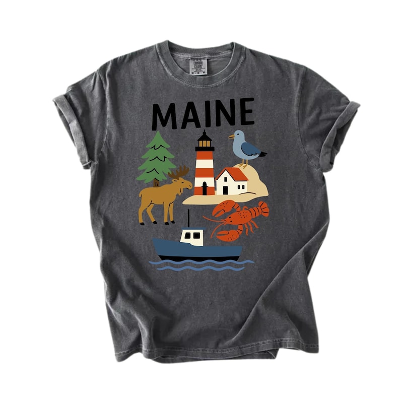May include: Dark grey t-shirt featuring the word "MAINE" above a graphic of a lighthouse, moose, lobster, boat, and seagull. The design is in red, orange, blue, green, and brown.