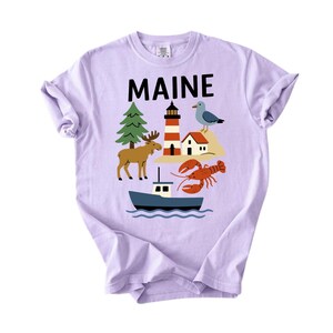 May include: A lilac t-shirt with the word "MAINE" in black block letters. The shirt features a colourful graphic of a lighthouse, moose, boat, lobster, and seagull, representing the state of Maine.