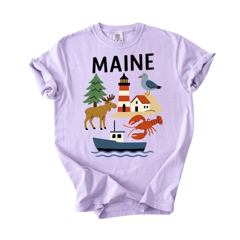 May include: A lilac t-shirt with the word "MAINE" in black block letters. The shirt features a colourful graphic of a lighthouse, moose, boat, lobster, and seagull, representing the state of Maine.