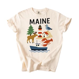 May include: Off-white t-shirt with a graphic of "MAINE" in black. The design includes a lighthouse, moose, lobster, boat, and seagull. The illustration is in a cartoon style, with a cream-colored background.