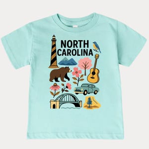 May include: A light blue t-shirt featuring a colorful graphic of North Carolina landmarks. The design includes a lighthouse, bear, mountains, flowers, a guitar, a car, and the text "NORTH CAROLINA".