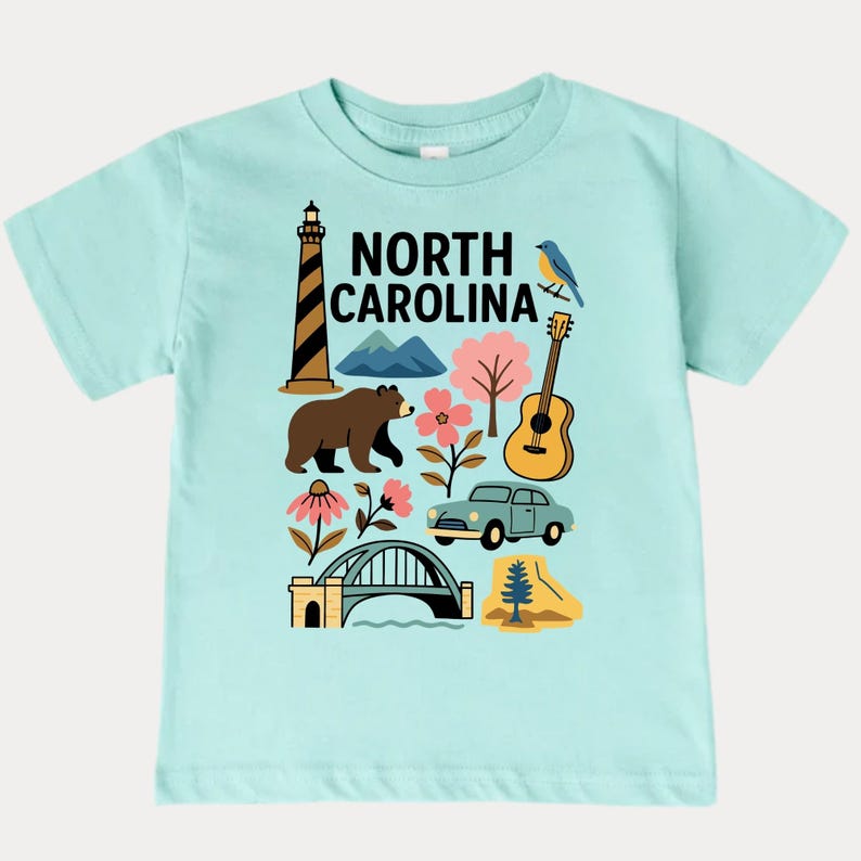 May include: A light blue t-shirt featuring a colorful graphic of North Carolina landmarks. The design includes a lighthouse, bear, mountains, flowers, a guitar, a car, and the text "NORTH CAROLINA".