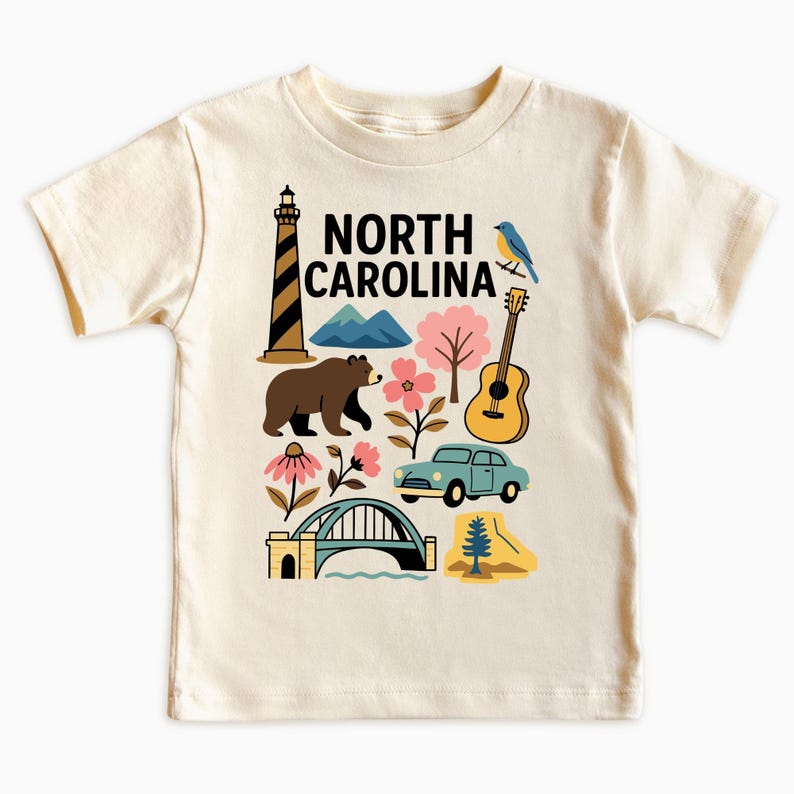 May include: Cream-colored t-shirt featuring a colorful graphic of North Carolina icons. The design includes a lighthouse, bear, mountains, flowers, a guitar, a car, a bridge, and the text "NORTH CAROLINA".