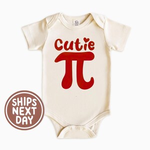 May include: Cream-colored baby onesie with short sleeves. The onesie features the red text "Cutie" above a red Pi symbol. The onesie has a snap closure at the bottom.