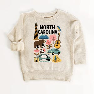 May include: A beige sweatshirt featuring a colorful graphic of North Carolina landmarks. The design includes a lighthouse, bear, mountains, flowers, a guitar, a car, a bridge, and the text "NORTH CAROLINA".