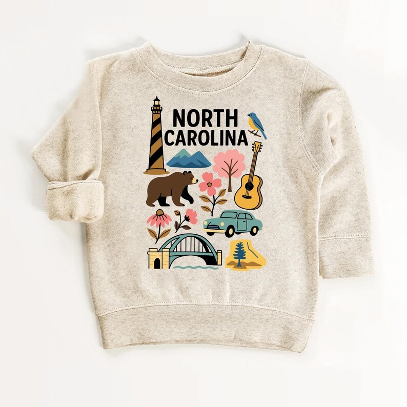May include: A beige sweatshirt featuring a colorful graphic of North Carolina landmarks. The design includes a lighthouse, bear, mountains, flowers, a guitar, a car, a bridge, and the text "NORTH CAROLINA".