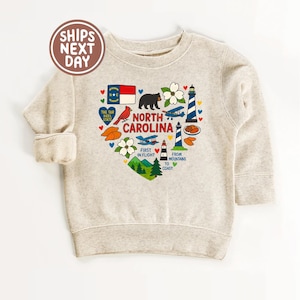 May include: A light beige sweatshirt featuring a heart-shaped design with North Carolina state symbols. The design includes a bear, cardinal, lighthouse, and the text "NORTH CAROLINA". The text "SHIPS NEXT DAY" is in a circle at the top.