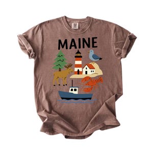 May include: A brown t-shirt displaying the word "MAINE" in black. The shirt features a graphic of a lighthouse, moose, lobster, boat, and other Maine-themed illustrations. The t-shirt is made of a soft, comfortable material.