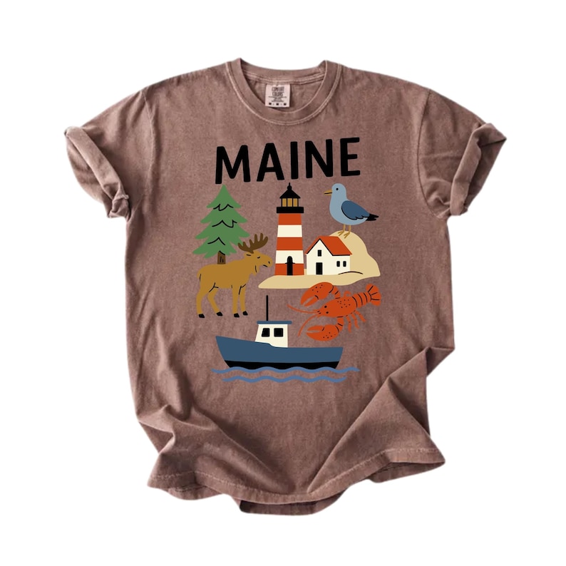 May include: A brown t-shirt displaying the word "MAINE" in black. The shirt features a graphic of a lighthouse, moose, lobster, boat, and other Maine-themed illustrations. The t-shirt is made of a soft, comfortable material.