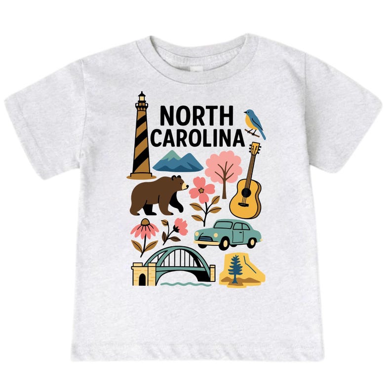May include: A light gray t-shirt featuring a colorful graphic of North Carolina landmarks. The design includes a lighthouse, bear, mountains, flowers, a guitar, a car, a bridge, and the text "NORTH CAROLINA".