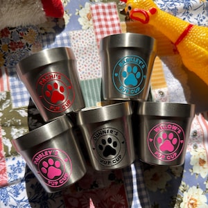 May include: Five stainless steel pup cups with colorful paw print designs. Each cup features a different name: Pachi's, Barkley's, Paisley's, Gunner's, and Willow's. The cups are arranged on a patchwork quilt with a rubber chicken toy.