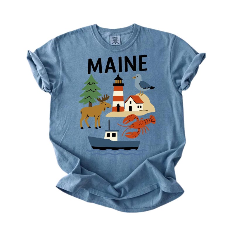 May include: Blue t-shirt with the word "MAINE" in black block letters. The shirt features a graphic of a lighthouse, moose, seagull, lobster, boat, and a small house. The shirt is made of a soft material.