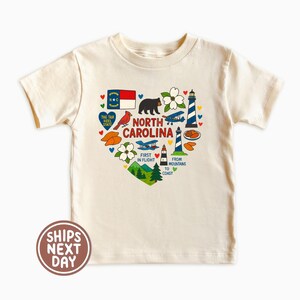 May include: Cream-colored t-shirt featuring a heart-shaped design with North Carolina state symbols. The design includes a bear, lighthouse, airplane, and the state flag. Text includes "North Carolina", "First in Flight", and "From Mountains to Coast".