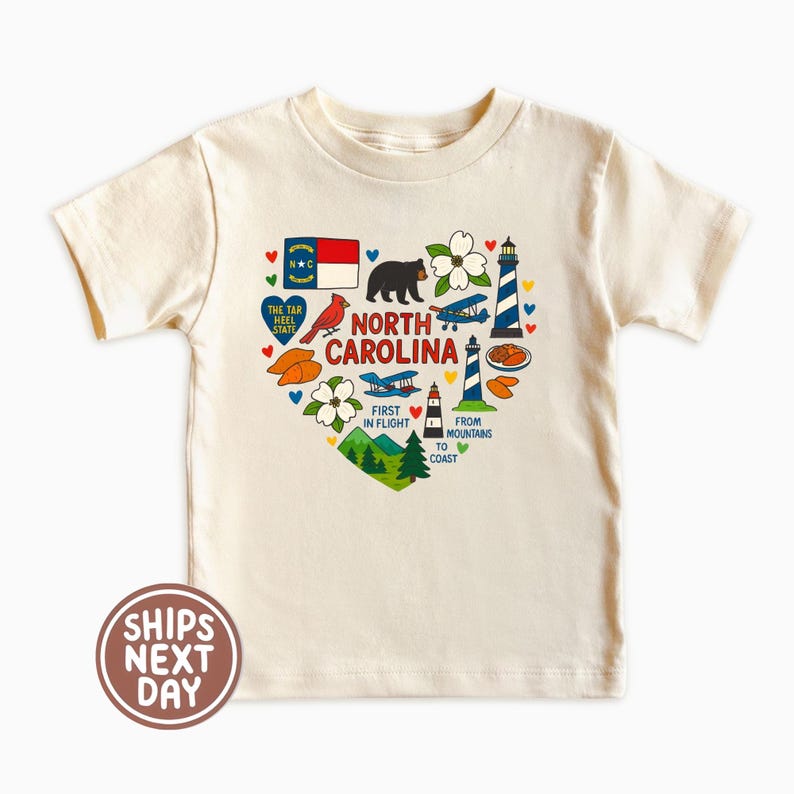 May include: Cream-colored t-shirt featuring a heart-shaped design with North Carolina state symbols. The design includes a bear, lighthouse, airplane, and the state flag. Text includes "North Carolina", "First in Flight", and "From Mountains to Coast".
