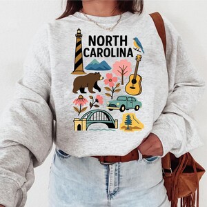 May include: A light gray sweatshirt featuring a colorful graphic of North Carolina landmarks and symbols. The design includes a lighthouse, bear, mountains, flowers, a guitar, a car, a bridge, and the text "NORTH CAROLINA".