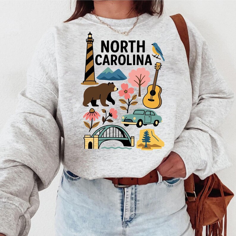 May include: A light gray sweatshirt featuring a colorful graphic of North Carolina landmarks and symbols. The design includes a lighthouse, bear, mountains, flowers, a guitar, a car, a bridge, and the text "NORTH CAROLINA".