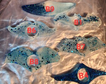 May include: Nine blue and white resin bookmark shapes with glitter embedded in the resin. The shapes are a bat, a mustache, a dolphin, a heart, a book, a bat, a heart, a dolphin, and a book. Each shape has a letter and number on it, for example, B1, B2, B3, etc.