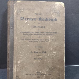 May include: An antique cookbook titled "Berner Kochbuch" with faded brown cover and text. The cover features ornate lettering and the publication year 1871. The book appears aged, with wear and tear visible on the edges.