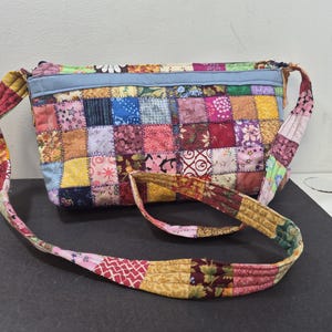 May include: A patchwork crossbody bag with a long strap. The bag is made of various fabric squares in a variety of colors and patterns, including floral, geometric, and solid colors. The strap is also made of patchwork fabric.