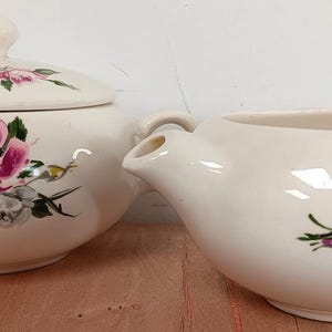 May include: A white ceramic sugar bowl and creamer set with a floral design featuring pink roses and green leaves. The set has a vintage look.