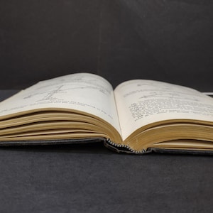 May include: An open book with aged, yellowed pages, revealing printed text and diagrams. The book's spine is visible, and the pages are slightly curved. The background is a dark gray surface.