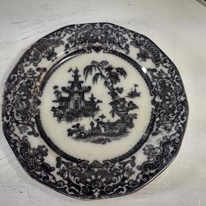 May include: A white ceramic plate with a black and white design. The design features a pagoda, trees, and other oriental motifs. The plate has a scalloped edge.