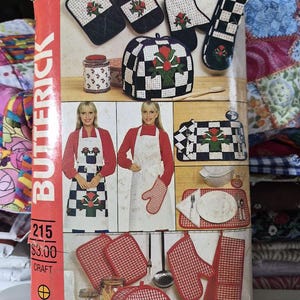 May include: A Butterick 215 craft sewing pattern envelope featuring illustrations of kitchen textiles including aprons, oven mitts, pot holders, and a tea cozy. The designs showcase black and white checkered patterns with red floral motifs and red gingham.