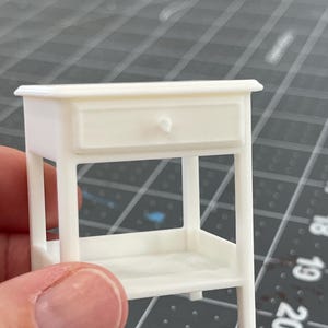 May include: A small, white miniature nightstand with a single drawer and a lower shelf. The nightstand has a curved drawer front and a small, round knob. The legs are straight and the overall design is simple and elegant. The nightstand is approximately 3 inches tall.