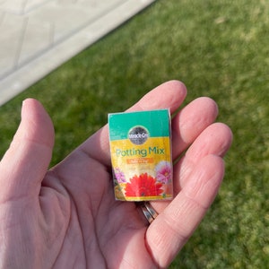 Peut inclure: Une petite boîte de terreau Miracle-Gro avec un fond jaune et une image de fleurs. La boîte est tenue dans une main.