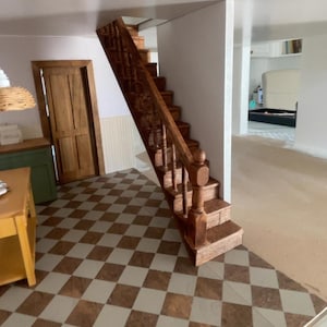 May include: Interior shot of a dollhouse with a staircase. The stairs are made of wood and have a dark brown finish. The floor is tiled in a brown and cream checkerboard pattern. The text "Victorias Farmhouse Dollhouse update! Stairs" is visible.