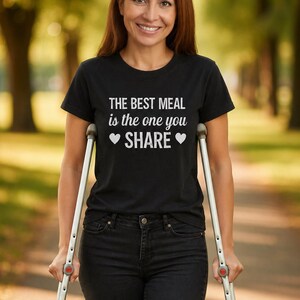 Peut inclure: Un t-shirt noir avec le texte "THE BEST MEAL is the one you SHARE" en blanc. La personne porte un jean noir et utilise des béquilles. L'arrière-plan est une scène extérieure floue avec des arbres.