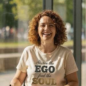 Peut inclure: Une femme en fauteuil roulant porte un t-shirt beige avec le texte "Starve the EGO feed the SOUL" en lettres noires et dorées. Elle sourit, a les cheveux roux bouclés et porte un jean bleu.