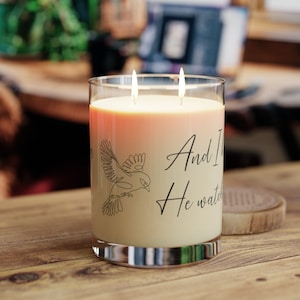 Puede incluir: Un tarro de vidrio para velas con un diseño degradado en color melocotón y blanco. La vela tiene dos mechas y presenta un dibujo de línea negra de un pájaro con el texto "And I knew He watched over me" en la parte delantera.