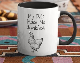 May include: White ceramic coffee mug with a black rim and handle. The mug has a black and white illustration of a chicken and the text "My Pets Make Me Breakfast".