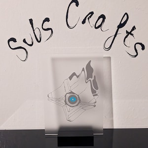 May include: A clear acrylic display stand with a black base, featuring a white and gray outline of a Ghost from the video game Destiny. The stand has the text "Subs Crafts" written on the wall behind it.