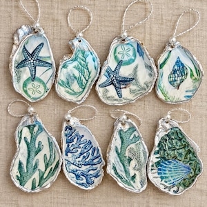 Könnte beinhalten: Acht dekorative Austernschalen-Anhänger mit Decoupage-Motiven von blauen Seesternen, grüner Koralle, Seeigeln und Muscheln. Jede Schale hat einen silberfarbenen Rand und eine weiße Kordelschlaufe mit einer kleinen weißen Perle zum Aufhängen.