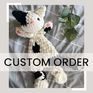 May include: A handmade crochet cat toy in cream, black, and pink. The cat has black patches on its ears and body, with pink accents on its paws and cheeks. The text "CUSTOM ORDER" is displayed across the image.