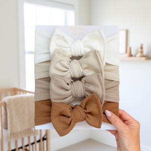 May include: A set of four waffle-knit headbands arranged vertically on a white display card labeled Harper + Remi. The headbands feature a knotted bow design and transition in color from cream to beige, light brown, and a warm medium brown shade.