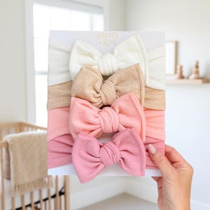 May include: A set of four fabric headbands on a white display card labeled Harper + Remi. The headbands feature a waffle-knit texture and a knotted bow design in a gradient of colors, including cream, tan, light peach, and dusty rose pink.