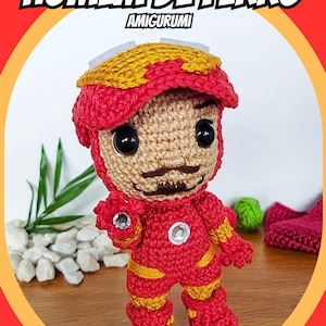 Pode incluir: Um brinquedo de amigurumi de crochê do Homem de Ferro, um super-herói da Marvel Comics. O brinquedo é vermelho e amarelo e tem um rosto detalhado com bigode. O brinquedo está em pé sobre uma superfície de madeira. O texto "HOMEM DE FERRO AMIGURUMI" está no topo da imagem.