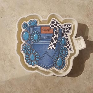 May include: A white mold featuring a denim pocket design with turquoise-colored gemstones and a black and white cow print bow. The word "Wrangler" is printed on a small brown label.