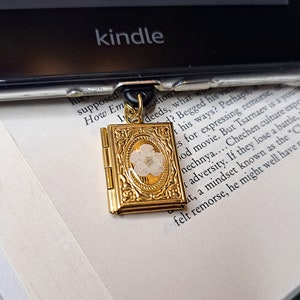 May include: A gold-colored book-shaped bookmark with a white pressed flower inside. The bookmark is attached to a black and gold chain.