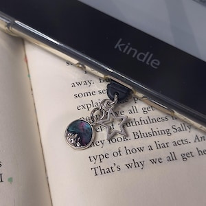 May include: A silver charm with a round, iridescent pendant and a star-shaped charm, attached to a Kindle e-reader. The e-reader is black with the word "kindle" printed on it. The charm is resting on the pages of a book.