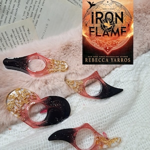 May include: Three black and pink resin bookmark rings with gold glitter accents. The rings are shaped like a teardrop with a hole in the center for a finger. The rings are on a white fuzzy surface. The book cover for "Iron Flame" by Rebecca Yarros is in the background.