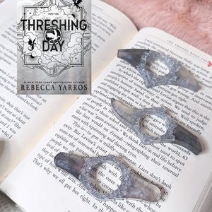 May include: A book titled Threshing Day by Rebecca Yarros with a cover featuring a dragon illustration and three grey, translucent, resin page holders with textured, silver-colored edges, each featuring a central circular opening for a thumb.