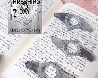 May include: A book titled Threshing Day by Rebecca Yarros with a cover featuring a dragon illustration and three grey, translucent, resin page holders with textured, silver-colored edges, each featuring a central circular opening for a thumb.