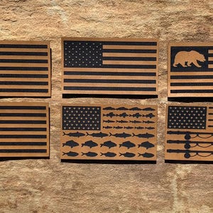 May include: A collection of six wooden American flag designs. Each flag features a unique design in the upper left corner, including a fish, bear, skull, and various fish and fishing rod silhouettes. The flags have a natural wood grain background with black stripes and details.