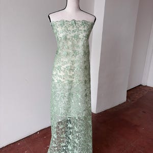 Op de afbeelding: Een lichtgroene kanten stof gedrapeerd over een mannequin. De stof heeft een bloemenpatroon en een geschulpte rand. Het kant is semi-transparant en onthult de mannequin eronder. De stof is vloerlang, geschikt voor jurken of andere kleding.