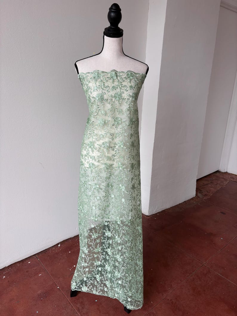Op de afbeelding: Een lichtgroene kanten stof gedrapeerd over een mannequin. De stof heeft een bloemenpatroon en een geschulpte rand. Het kant is semi-transparant en onthult de mannequin eronder. De stof is vloerlang, geschikt voor jurken of andere kleding.